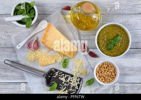 Leckere hausgemachte grüne Soße pesto mit Basilikum, Käse und Pinienkernen in Weiß Schüssel auf Holztisch mit Raspel, Mörtel, Basilikum, Knoblauch und oliv Stockfoto