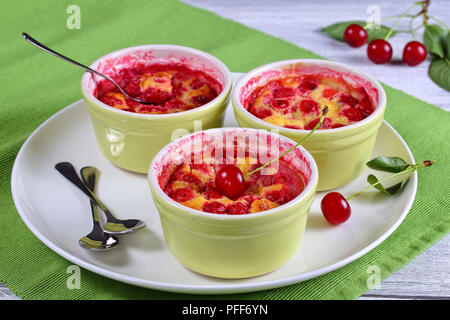 Leckere Sour Cherry clafoutis - klassische französische süße Frucht Vanille hausgemachten Dessert in Keramik Förmchen mit Löffeln auf Teller auf Holztisch, authent Stockfoto