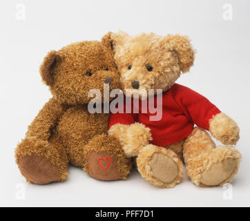 Zwei Teddybären nebeneinander sitzen gegeneinander gelehnt, einer trägt einen roten Pullover, aus der Nähe. Stockfoto