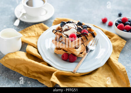 Belgische Waffeln mit Vanilleeis und Beeren beträufelt mit Schokolade und Kaffee im Hintergrund. Selektiver Fokus Stockfoto