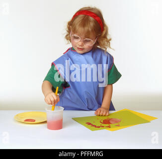 Mädchen Tragen einer Brille, Schürze und Kopfband, Pinsel in Plastik Tasse rote Wasser, rote Farbe auf die Platte und auf Stück farbiges Papier Stockfoto