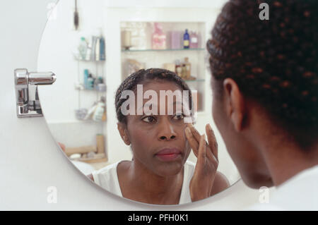 Reife Frau im weißen Hemd runder Spiegel suchen, zwei Finger auf die Wange, Regale mit Flaschen hinter sich. Stockfoto