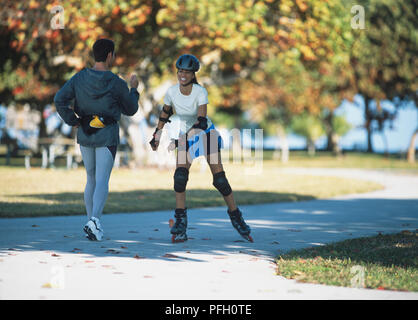 Mann in Nylon Jacke, Leggings, Power Walking Vergangenheit lächelnde Frau auf Inline-skates mit Knie Pads, Ellbogen Pads, Handgelenksschoner, Helm, auf Weg mit Bäumen. Stockfoto