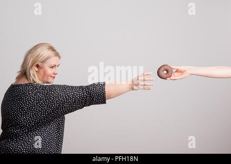 Ein glückliches übergewichtige Frau im Studio, für einen Donut, indem eine Hand eine nicht erkennbare Person gehalten wird. Stockfoto