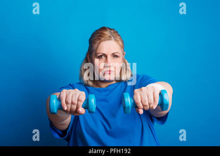 Porträt einer schweren übergewichtigen Frau mit Hanteln in den Händen. Studio shot auf blauem Hintergrund. Stockfoto