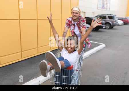Bild der junge lächelnde Freunde über gelbe Wand. Viel Spaß mit Einkaufswagen. Stockfoto