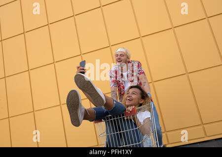 Bild der junge lächelnde Freunde über gelbe Wand. Viel Spaß mit Einkaufswagen und selfie machen. Stockfoto