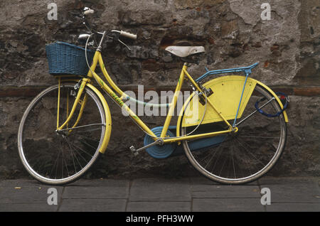 Die gelben Frauen Fahrrad mit einem Weidenkorb auf Vorderrad montiert, lehnte sich gegen eine verwitterte alte Mauer Stockfoto