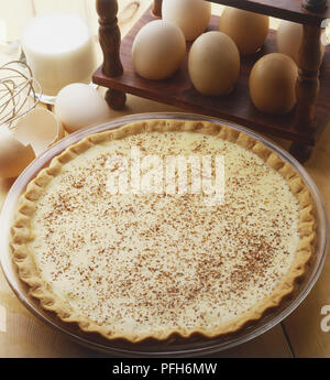 Ein custard Pie bestreut mit Puderzucker Schokolade, neben frische Eier in hölzernen Ei Fach, zerbrochene Eierschale und einem Schneebesen, Nahaufnahme, hohe Betrachtungswinkel Stockfoto