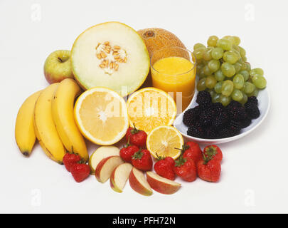 Auswahl an frischem Obst, einschließlich Melone ganz und halbiert, Bananen, Orangen- und Zitronenbäumen, Apfelscheiben, Erdbeeren, Trauben und Brombeeren, und ein Glas Orangensaft Stockfoto