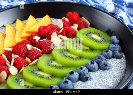 Gesundes Frühstück - lecker Chia Samen Pudding mit Himbeeren, Blaubeeren, Pfirsiche und Kiwis Scheiben in schwarz Schüssel auf alten Holztisch, selektive Stockfoto
