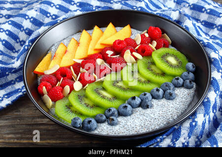 Köstliche Chia Samen Pudding mit Himbeeren, Blaubeeren, Pfirsiche und Kiwis Scheiben in schwarz Schüssel auf alten Holztisch, horizontale Ansicht von oben, c Stockfoto