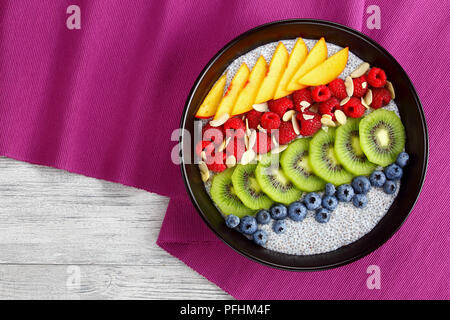 Köstliche Chia Samen Pudding mit Himbeere, Heidelbeere, Pfirsich und Kiwi Scheiben bestreut mit mandelflocken in schwarz Schüssel auf dem Tisch Matte auf alten Woo Stockfoto