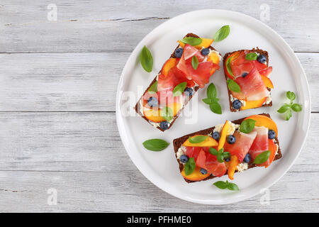 Leckere bunte Sandwiches mit Frischkäse, Blaubeeren, Pfirsich, Schinken Scheiben und Blätter Basilikum auf Roggenbrot auf weiße Platte, auf hölzernen Tisch, Stockfoto