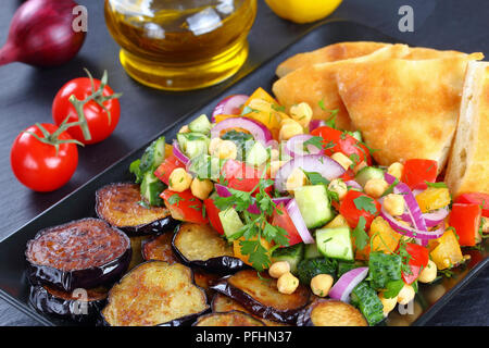 Köstliche mediterrane Kichererbsen Salat mit Gurken, Tomaten, roten Zwiebeln und gebratenen Auberginen auf schwarzem Teller mit Käse gefüllte Fladenbrot, auf dunklen Holz Stockfoto