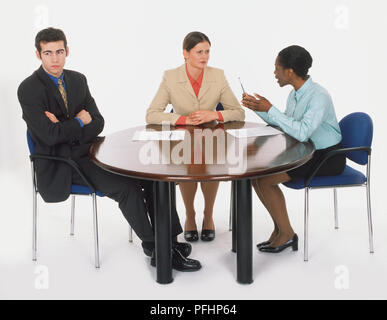 Drei Menschen in Anzügen am runden Tisch sitzen, Mann mit verschränkten Armen und seitwärts, als zwei Frauen eine Debatte haben. Stockfoto