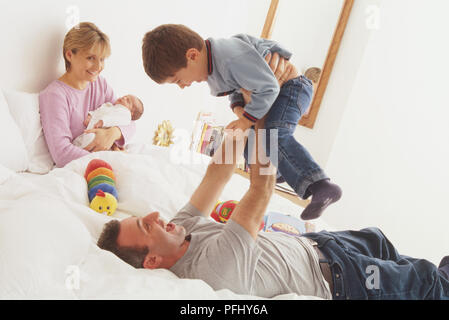 Lächelnde Frau im Bett sitzen Halten eines neugeborenen Baby, Mann zurück auf dem Bett liegend Heben ein Kleinkind in der Luft, von der Seite. Stockfoto
