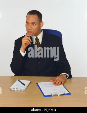 Mann in Anzug an einem Schreibtisch mit seiner Hand auf seinem Kinn, Kugelschreiber, Notizblock und Zwischenablage vor Ihm sitzt, Vorderansicht. Stockfoto