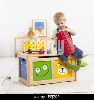 Junge versucht, Red Box mit Red Box zu öffnen, während Sitzen auf Holz- trunk nächsten Doppeldeckerbus, Regal- und Teddybären im Hintergrund zu verdoppeln, verschwommen. Stockfoto