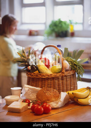 Auswahl der Milchprodukte, Tomaten, Bananen, Laden von Brot und Korbwaren Korb mit frischem Obst und Gemüse Küche Arbeitsplatte gefüllt, Frau waschen Geschirr in der Spüle im Hintergrund. Stockfoto