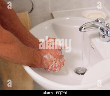 Mann, der über dem Waschbecken, Tippen läuft, reibt die Hände zusammen, in schäumende Seife, Nahaufnahme, Handtuch, Seifenschale und Bad Fliesen im Hintergrund Stockfoto