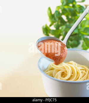 Schöpfkelle mit Tomatensoße über Schüssel Spaghetti, frischem Basilikum im Hintergrund Stockfoto
