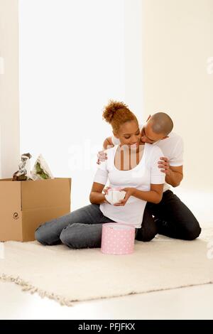 Junge Frau sitzt auf dem Boden Eröffnung anwesend, junger Mann hinter ihr und küsste sie auf den Hals Stockfoto