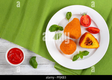 Gebratene arancini-risotto Kegel gefüllt mit Fleisch Ragu, und grünen Erbsen auf weiße Platte mit Tomaten und Basilikum. Tomatensauce in Soße auf Tisch, aut Stockfoto