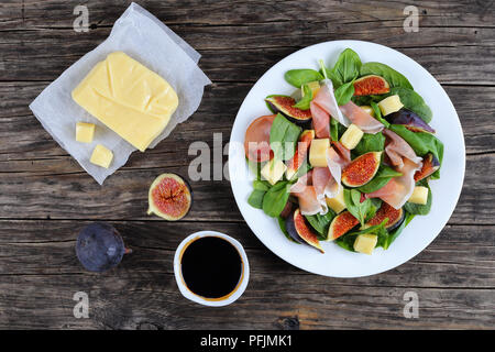 Spinat, Feigen, dünn geschnittene italienischer Schinken und Mozzarella Salat auf weiße Platte auf dunklen, hölzernen Küchentisch mit Stück Käse auf dem Papier und Bal Stockfoto