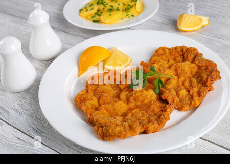 Köstliche golden braun klassisches Wiener Schnitzel- oder paniertes Schnitzel serviert auf weiße Platte mit Zitronenscheiben auf dunklen Holztisch mit Glas r Stockfoto