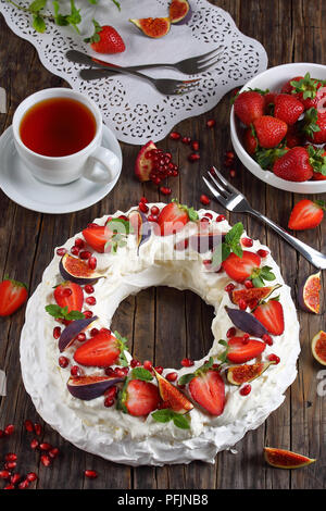 Pavlova kuchen Kranz der französischen Baiser und Sahne, dekoriert mit Erdbeere, Feigen, Granatapfel und Minze auf dunklen Tisch mit Tasse Tee und Ingr Stockfoto