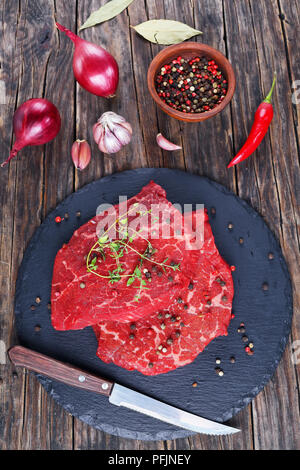 Zwei Marmorierte Rindfleisch steaks Ausgegossen über mit Öl und garniert mit frischem Grün Thymian auf schwarzem Schiefer Fach mit Messer, Gewürze, Zwiebel, Lorbeer auf alten Stockfoto