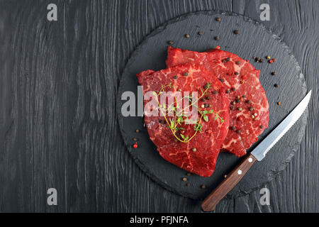 Zwei Rohmaterial Marmorierte Rindfleisch Steaks mit frischem Grün Thymian auf schwarzem Schiefer Fach mit Messer und Mischung der pfefferkörner auf schwarzen Holztisch überstieg, Horizont Stockfoto
