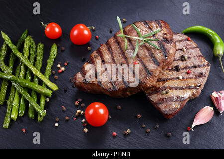 Hot köstliche gegrillte Rindersteaks mit Streifen mit Tomaten serviert gegrillter Spargel, Knoblauch und Gewürzen auf schwarzem Schiefer Platte, horizontale Ansicht aus Stockfoto