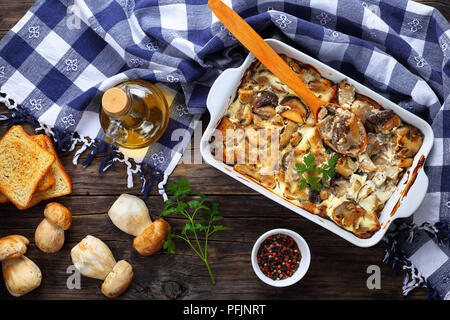 In Scheiben geschnittenen Champignons mit saurer Sahne, Zwiebel und Butter in der auflaufform auf alten hölzernen Tisch gekocht, eine Flasche Olivenöl, rohe Pilze, geröstetes Brot auf Ba Stockfoto