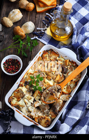 Köstlich in Scheiben geschnittenen Champignons mit saurer Sahne, Zwiebel und Butter in der auflaufform auf alten hölzernen Tisch gekocht, eine Flasche Olivenöl, rohe Pilze, geröstetes b Stockfoto