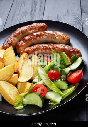 Leckere heiße gegrillte Würstchen, gebackene Kartoffeln und frischem Gemüse romaine Kopfsalat Salat auf schwarze Platte auf schwarzen Brettern, Seite, Ansicht von oben, Stockfoto