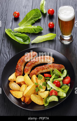 Leckere heiße gegrillte Bratwurst, gebackene Kartoffeln und frischem Gemüse romaine Kopfsalat Salat auf schwarze Platte auf schwarzer Holztisch mit Glas frisch werden. Stockfoto