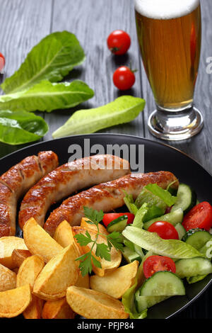 Leckere heiße gegrillte Bratwurst, gebackene Kartoffeln und frischem Gemüse romaine Kopfsalat Salat auf schwarze Platte auf schwarzer Holztisch mit Glas frisch werden. Stockfoto
