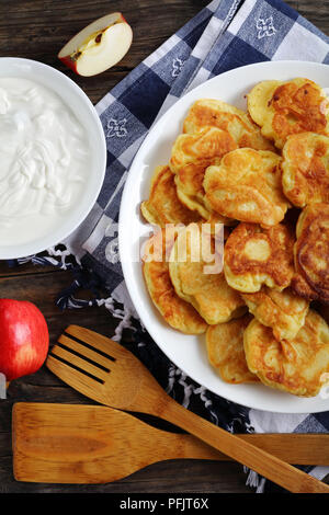 Leckere Pfannkuchen mit saftigen Stücke von Apple geladen, auf weißem Schild auf alten, dunklen Holztisch mit saurer Sahne in der Schüssel und Äpfel im Hintergrund, vertic Stockfoto
