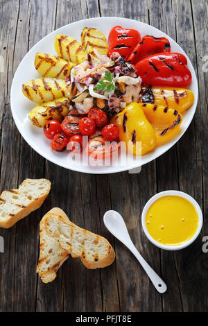 Leckere gemischte Meeresfrüchte Salat serviert auf weiße Platte mit gegrilltem Gemüse. basic rue Sauce und geröstetem Baguette auf Holztisch, vertikale Ansicht von Stockfoto