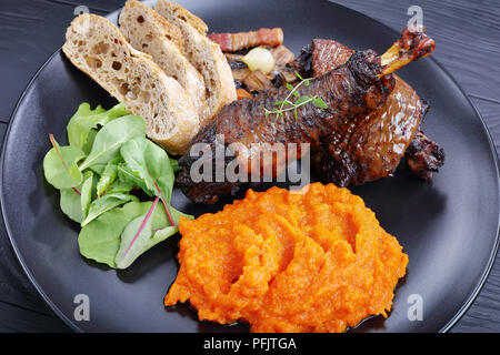 Ein Teil von Coq au Vin - klassische französische Hühnereintopf auf schwarze Platte mit Mix aus salatblättern serviert, Kürbispüree und Brot, horizontale Ansicht aus Stockfoto