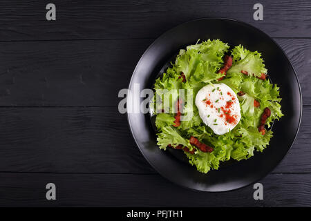 Traditionelle französische Salat Lyonnaise mit frisee Eisbergsalat, Speck und ein pochiertes Ei auf schwarzem Teller auf Holztisch, einfach und Gesundes Rezept, Ansicht von Abo Stockfoto