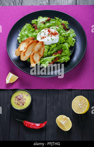 Klassische französische Salat Lyonnaise mit frisee Eisbergsalat, heißem Speck und einem frisch pochiertes Ei auf schwarze Platte, Vinaigrette Dressing in kleinen Schüssel am backgrou Stockfoto