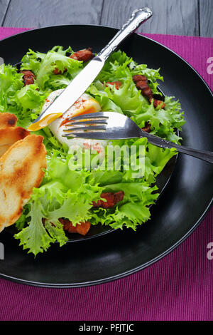 Frisee Eisbergsalat, knusprigem Speck und einem pochierten Ei Salat - frische und köstliche Lyonnaise Salat, auf schwarzem Teller serviert mit gerösteten Baguette, authentische recip Stockfoto