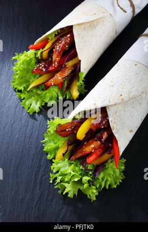 Tortilla gerollte um eine Füllung - rotkohl Salat, Paprika Streifen und Fried Chicken Fleisch auf schwarzer Schiefer Fach, auf Holztisch, Ansicht von abov Stockfoto