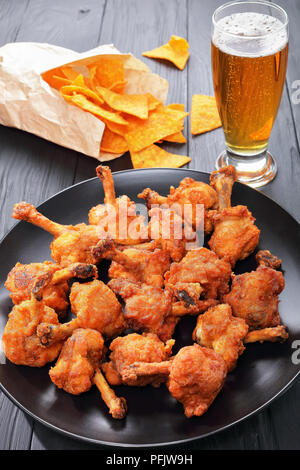 Leckere frittierte knusperiges Huhn Winglets mit freiliegenden Knochen auf eine schwarze Platte auf dunklen Holztisch zerschlagen. Glas frisches Bier und Nachos in Pape Stockfoto