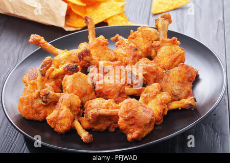 Leckere frittierte knusperiges Huhn Winglets mit freiliegenden Knochen auf eine schwarze Platte auf schwarzen Holztisch zerschlagen. nachos in Papiertüte im Hintergrund, vi Stockfoto