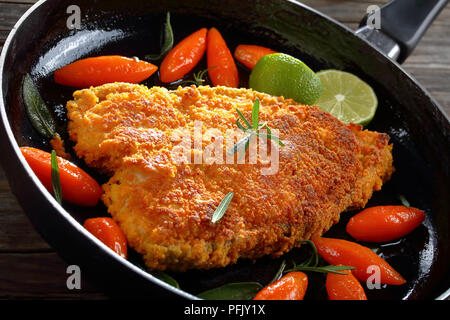 Gebratene panierte weißen Fisch Steak mit glasierten Karotten, schneiden Sie in Tourne (ovale Form), frische Kalk Schichten, Salbei und Rosmarin in Skillet auf Holztisch, vie Stockfoto