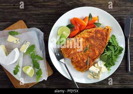 Lecker gebratene Panierte weißen Fisch Steak serviert auf weiße Platte mit glasierten Karotten, frische Kalk Schichten, blau Schimmelkäse und Rucola, Zutaten Stockfoto